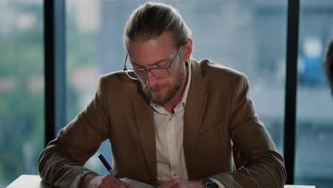 a young blond man with glasses, an office worker, sits at a table in a light suit and with a watch on his hands and thinks about solving issues. thinking about your work in a modern office with a panoramic window