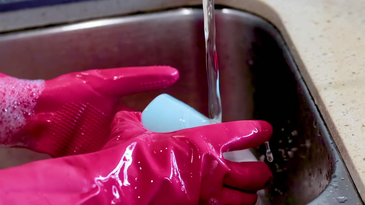 Close-up of pink-gloved hands rinsing a blue cup under running water in a kitchen sink.