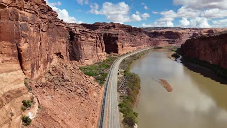 aerial drone shot of highway in desert, railroad tracks, tall vibrant red sandstone cliffs, colorado river, potash road in moab utah, blue sky with clouds,