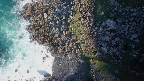 Aerial-overhead-shot-of-surf-breaking-on-wild,-rocky-coast-on-a-sunny-day