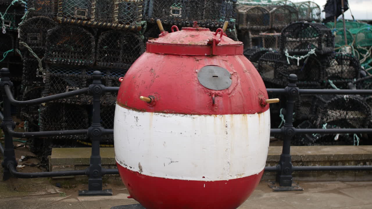 A red and white buoy sitting on a paved area near stacked fishing pots, surrounded by black metal railings in Scarborough, North Yorkshire in England