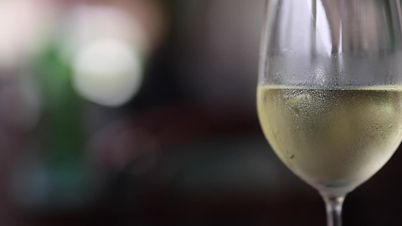 Una mano tiene in mano un bicchiere di vino bianco ricoperto di condensa su uno sfondo bokeh leggero