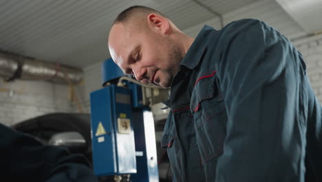 close-up of a focused mechanic in a blue uniform examining something off-camera in an automotive workshop with blurred background equipment