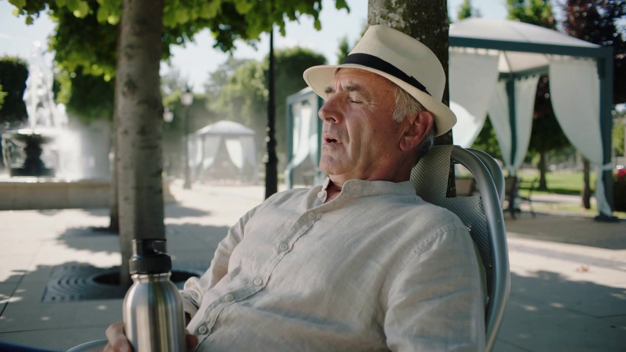 Hombre mayor relajado sosteniendo una botella de acero con sombrero de paja, cerrando los ojos mientras la luz del sol se filtra en la plaza.