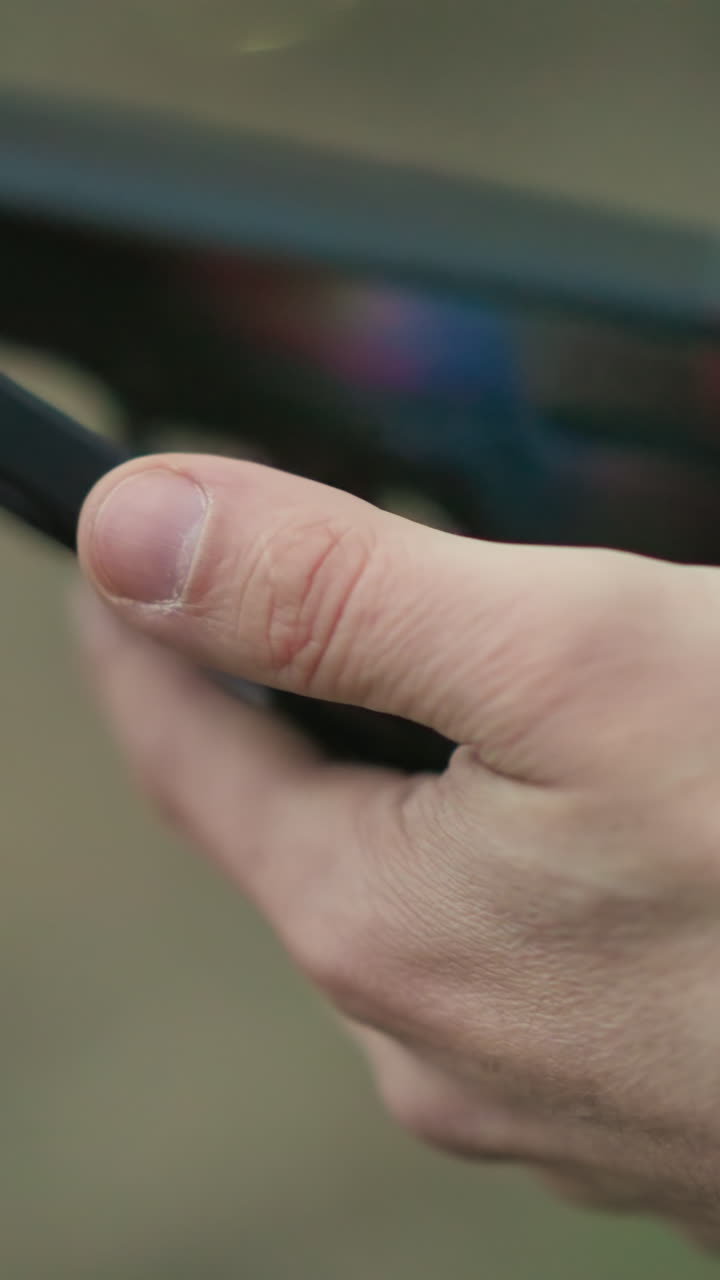 una vista de cerca de un hombre con una chaqueta verde sosteniendo un teléfono celular, operándolo y un arma, centrándose en sus manos