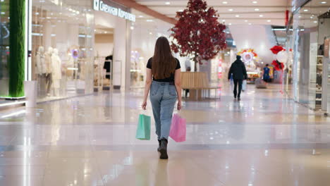 back view of a woman in jeans carrying shopping bags while walking in a modern, well-lit mall, with other shoppers ahead and light reflecting on the tiles