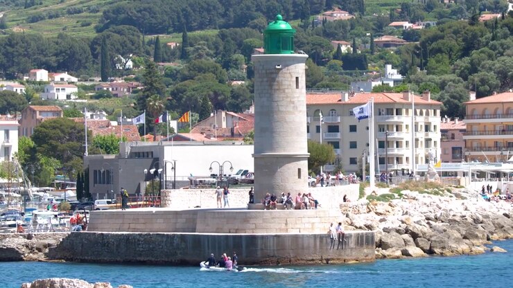 Ampia inquadratura fissa di un faro in pietra che si affaccia su un porto mediterraneo e un villaggio costiero