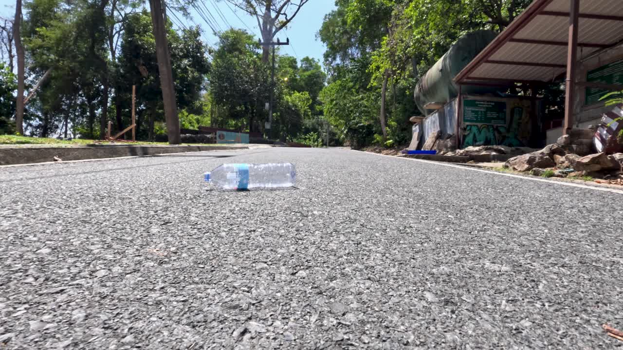 Empty plastic bottle rolls across sunlit asphalt road, low angle, outdoor, midday, urban environment