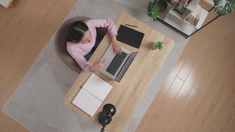 vista superior zoom out toma de una mujer cansada que tiene dolor de cabeza escribiendo en una computadora portátil mientras está sentada en el escritorio de madera en casa