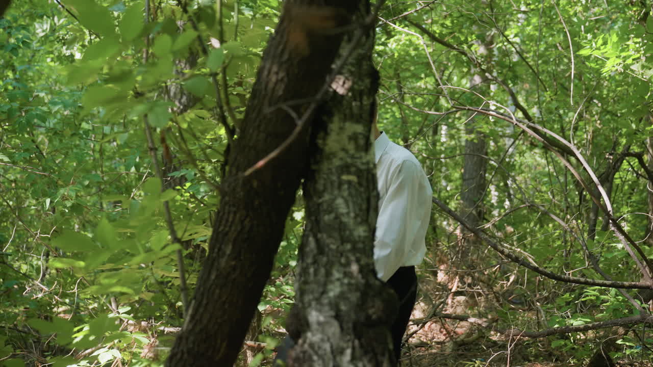 Young man in white shirt walking carefully through dense forest carrying bag, navigating branches and foliage under daylight, creating atmosphere of exploration and cautious movement