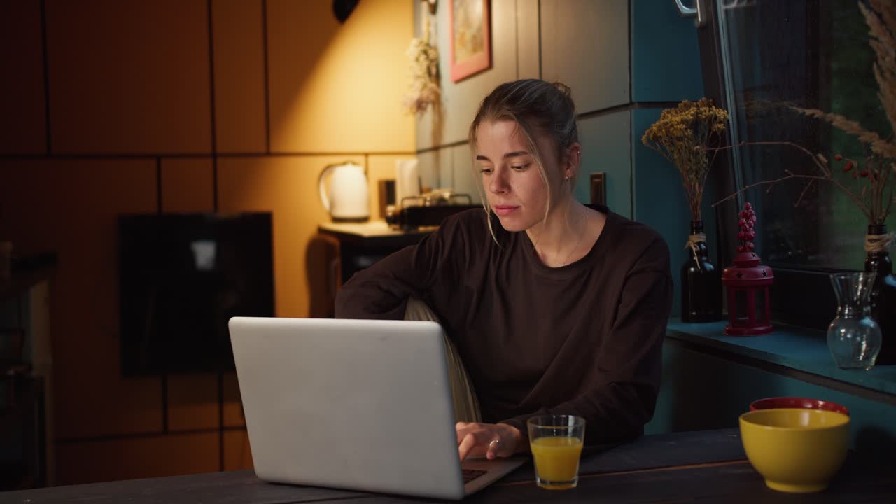 A happy blonde girl rejoices and looks at the screen of a white laptop in a dark evening room illuminated by a yellow lamp