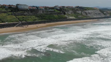 many surfers enjoying waves at basque coast beach, people relax on sand, green cliffs of biarritz, france. aerial forward