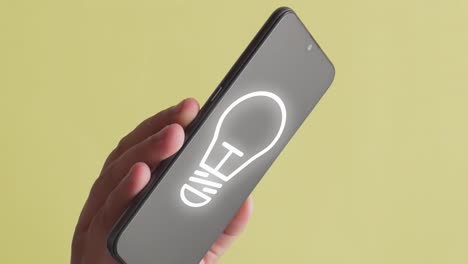 Hand-holding-a-mobile-phone-where-the-animation-of-symbol-of-a-light-bulb-or-idea-appears,-yellow-background