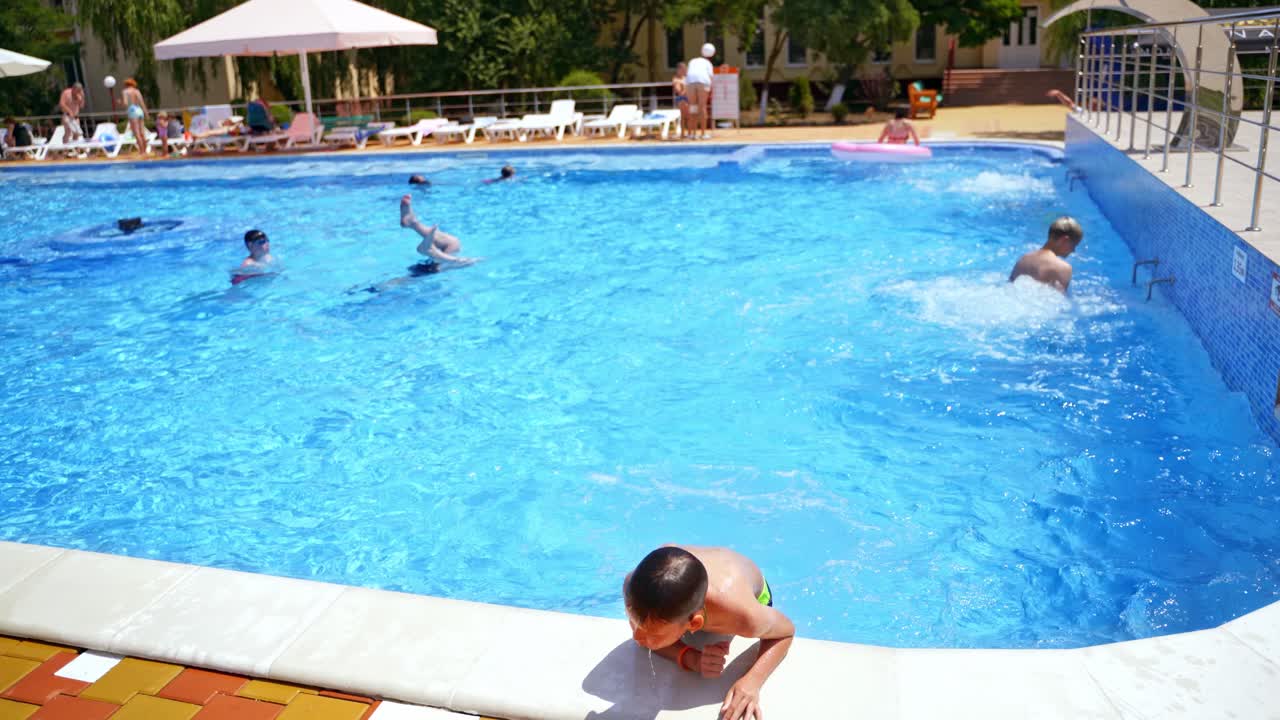 Children in the open swimming pool. Teenage boy jumping into the blue water of the pool in summer time. Happy vacation in water.