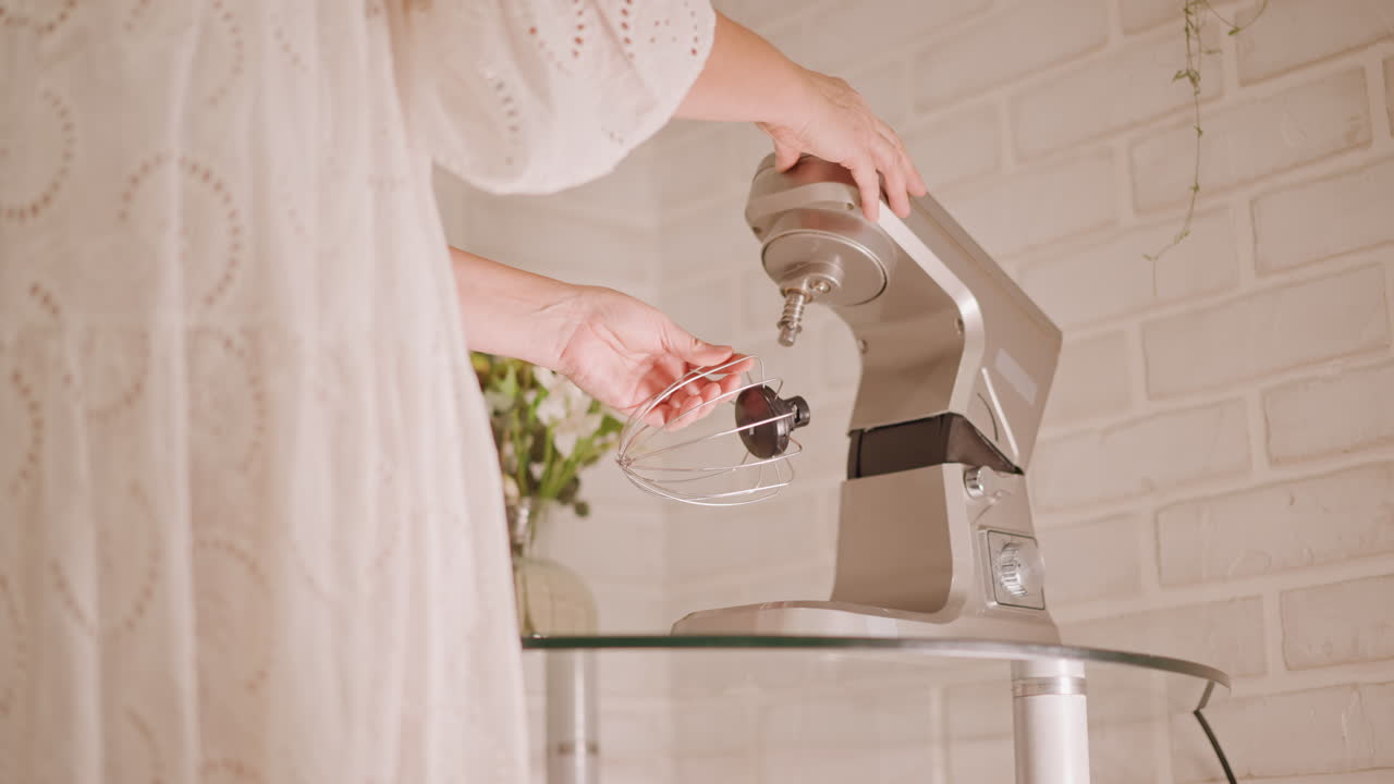 Hands attach beater to mixer on glass table by white brick wall, vase with flowers and tray of baked goods visible, warm soft light, home baker preparing pie crust and ingredients with calm ritual