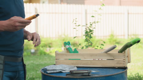 side view gardener removing tools, dropping on blue drum, dusting hand, apron belt on waist, wooden crate with green handles, gloves visible, yard house fence background
