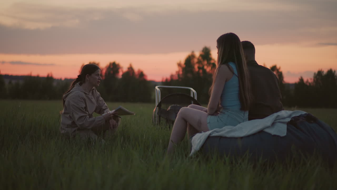 close up of woman crouching on grass holding notebook and pen interacting with young adult sitting on cushion listening during sunset field session