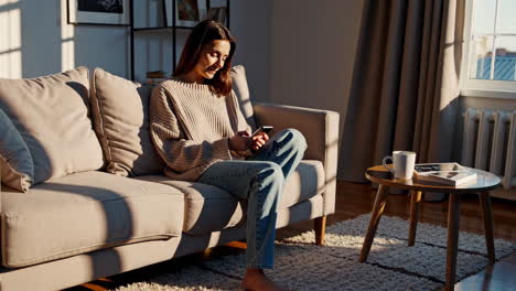 woman relaxing on couch while using smartphone in sunny living room