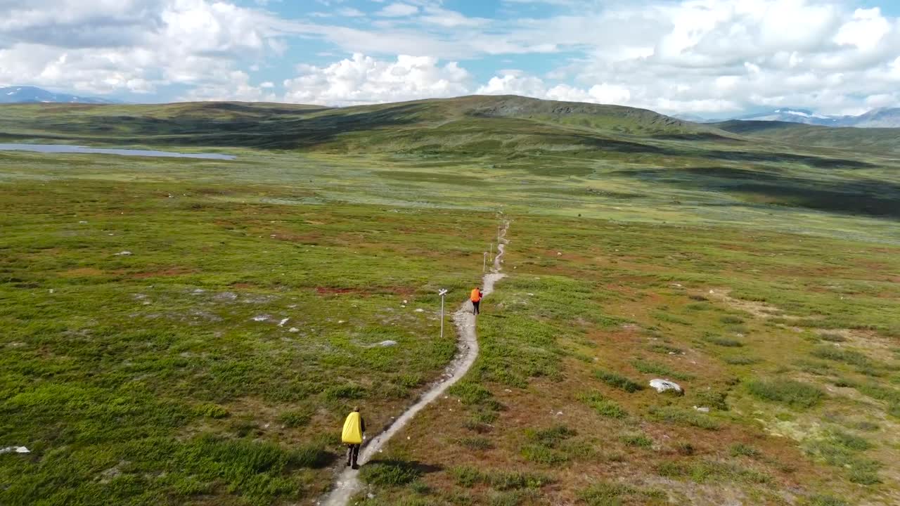 Aerial drone footage following and flying behind a couple or to hikers walking on a rural small muddy brown road on sweden grassy green and brown lanscapes during cloudy day. Hills visible on horizon.