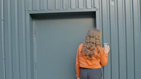 brown haired woman in orange jacket walks towards urban building, pushes key code, rear view