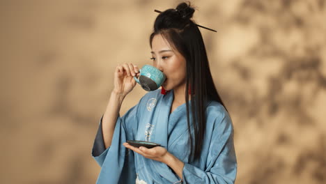 mujer joven asiática en kimono azul bebiendo té de una taza de cerámica y sonriendo a la cámara