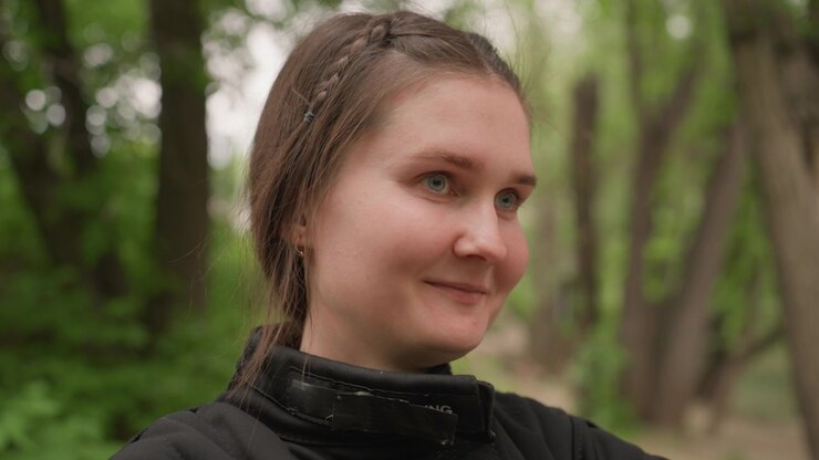 Primo piano di giovane donna con capelli intrecciati che indossa abbigliamento outdoor nero, in piedi in un ambiente naturale. La donna rimane per lo più immobile, girando occasionalmente la testa per guardare.