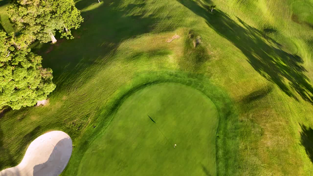 Drone captures a golf course with bunkers and green under golden hour lighting, highlighting shadows and landscape contours