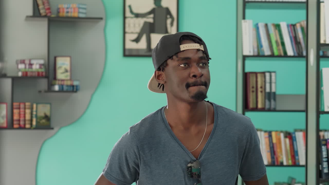 Young man in casual t-shirt and cap dances playfully in study room surrounded by colorful bookshelves and framed wall art, exuding energy and confidence against vibrant teal walls