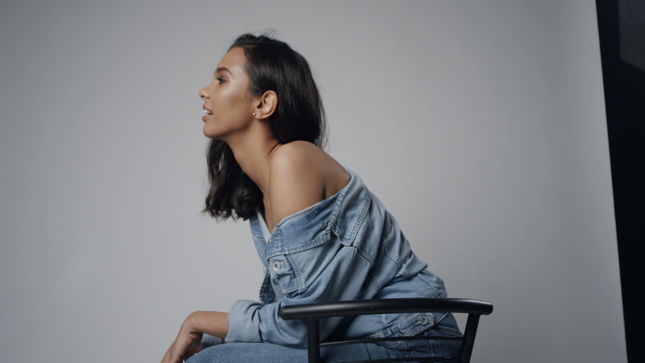 Portrait of female model posing to the camera while sitting on the chair on the white wall in the photostudio