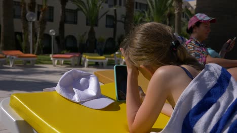 girl teenager lying on chaise longue near swimming pool and using mobile phone