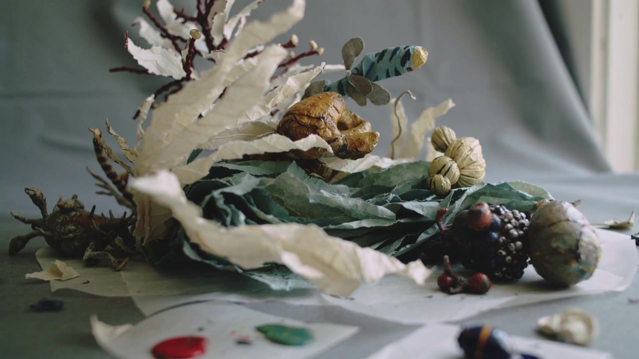 Pulling camera to reveal paper leaves and sculpted fruit on tabletop by window, with paint puddles