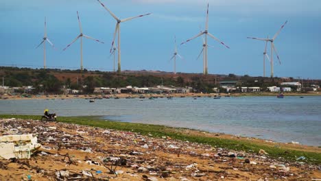 rotating blades of clean and renewable wind power farm in background and polluted beach shore with plastic,garbage and dirt