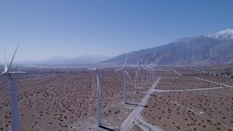 Drohne-Verfolgt-Windmühlen-In-Der-Wüste-Und-Enthüllt-Eine-Weitere,-Mit-Schneebedeckten-Bergen-Im-Hintergrund