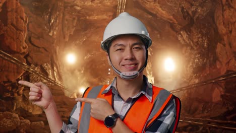 close up of asian male engineer with safety helmet smiling and pointing to side while standing in underground mine tunnel