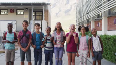 Diverse-group-of-schoolchildren-wearing-backpacks-smiling-and-standing-in-a-row-at-school-yard