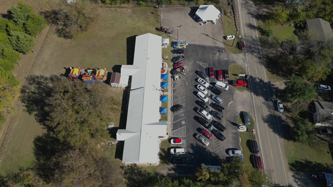Órbita suave de drone em 4K revela um evento comunitário ao lado de um grande edifício, com castelos infláveis coloridos, carros estacionados, gramados abertos e árvores ao redor
