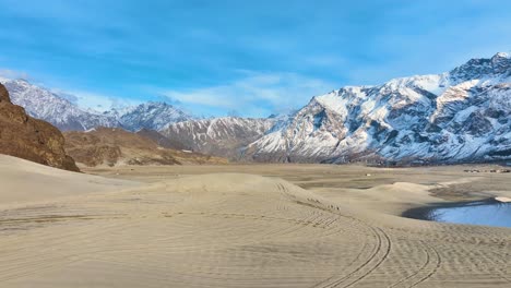 Luftaufnahme-Einer-Malerischen-Landschaft-Mit-Einer-Sandwüste-Im-Vordergrund-Mit-Reifenspuren-Und-Schneebedeckten-Bergen-Unter-Einem-Blauen-Himmel-Im-Hintergrund