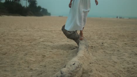 Una-Mujer-Vestida-De-Blanco-Camina-Sobre-Un-Tronco-En-La-Playa-Durante-La-Puesta-De-Sol,-Capturando-Un-Momento-Sereno-De-Soledad