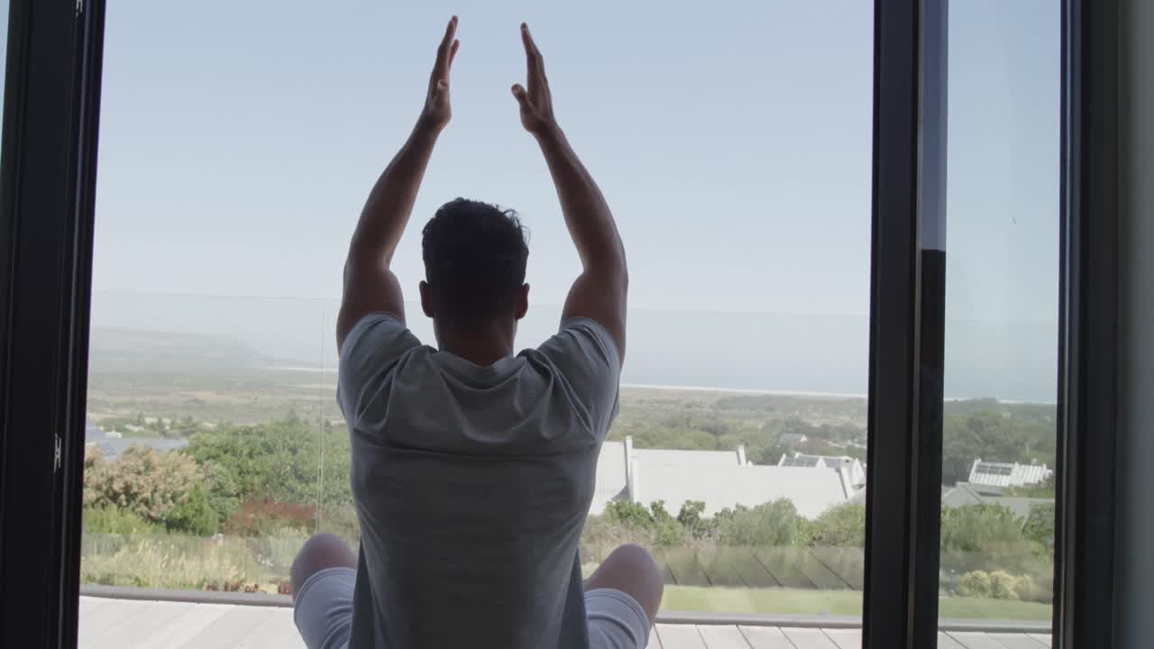 vista trasera de un hombre biracial sentado en un balcón soleado practicando meditación de yoga, cámara lenta