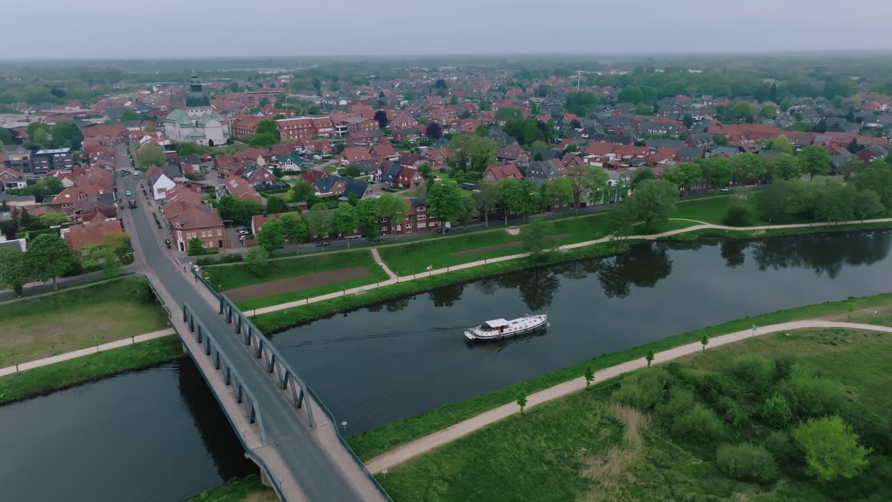 Aerial zoom in of a barge crossing beneath a bridge in a small German town. Overcast skies and calm water add to the serene atmosphere.