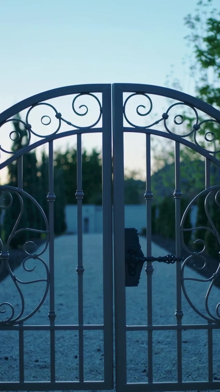Vertical video: Zooming camera focusing on ornate wrought iron gate at dusk, showing locked chain