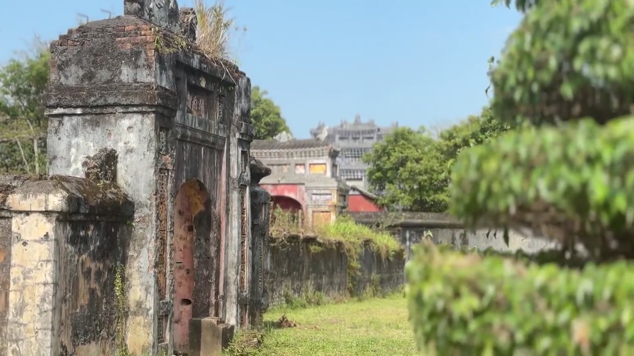 Pemandangan dekat topiary bertingkat yang dipangkas rapi dalam pot dekoratif, dengan arsitektur batu bersejarah dan area taman di dalam Kota Kekaisaran di Hue, Vietnam.
