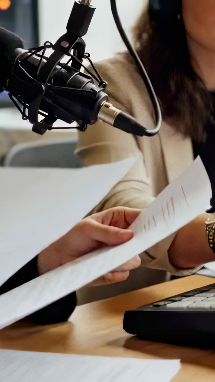 Vertical video: Taking pages, host in beige blazer preparing to read into mic at desk, mixer