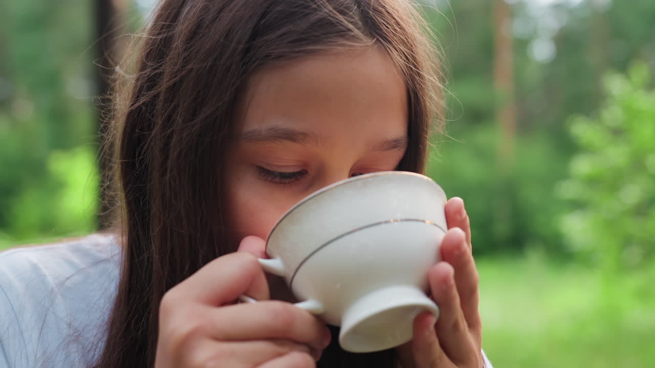 밝은 초록색 자연으로 둘러싸인 숲에서 평화로운 가족 피크닉을 즐기며 흰색 컵에 담긴 차를 마시는 소녀, 편안한 표정으로 따뜻한 음료를 즐기고, 부드러운 햇빛이 나무 사이로 스며들고 있어요.