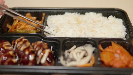 chopsticks picking up rice from dosirak or korean lunchbox with cutlets and kimchi - close-up