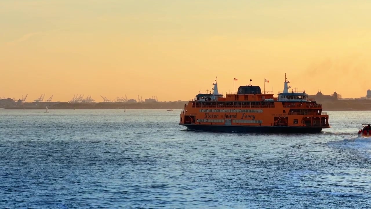 New York, ABD, 26 Mayıs 2026: New York Limanı'ndaki Staten Island Feribotu üzerinde altın saat ışığı. Sakin mavi su üzerinde, uzakta vinçler ve yelkenlilerle birlikte turuncu bir geminin geniş görünümü.
