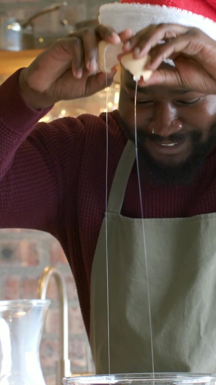 Video verticale: Rompendo le uova, uomo con cappello da Babbo Natale e grembiule che prepara l'impasto festivo mentre si asciuga il viso, ciotola