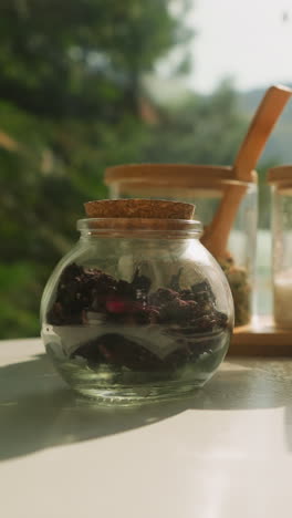 mesa muestra componentes para té de hierbas. recipientes de vidrio llenos de flores y hierbas secas contra la ventana con vista a la naturaleza. bebida natural