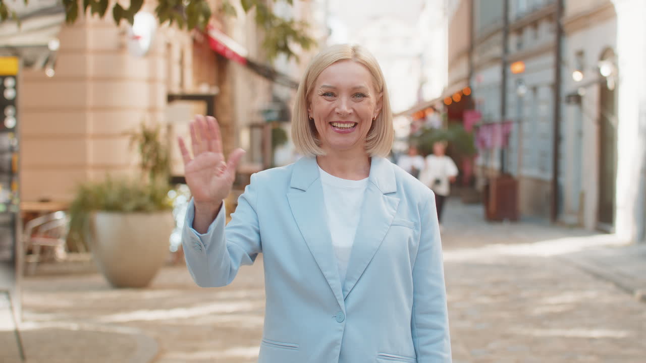 Happy mature businesswoman looking at camera waving hello greeting gesture friendly on city street