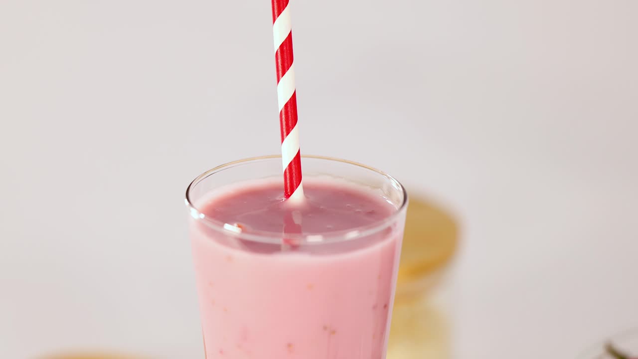 Batido rosa en vaso transparente, pajita a rayas, iluminación brillante, movimiento lento de la cámara hacia arriba, fondo minimalista